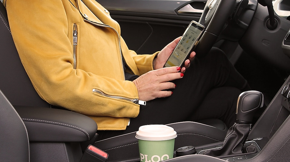 En kvinna med gul jacka som sitter i en bil och använder St1-appen Fuelpay för att välja pump och tanka med mobilen hon dricker kaffe från PLOQ.